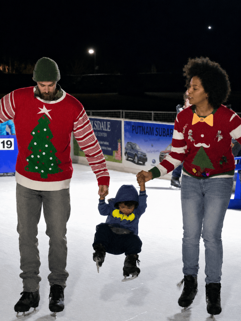 Two adults in holiday sweaters hold a child's hands while ice skating.