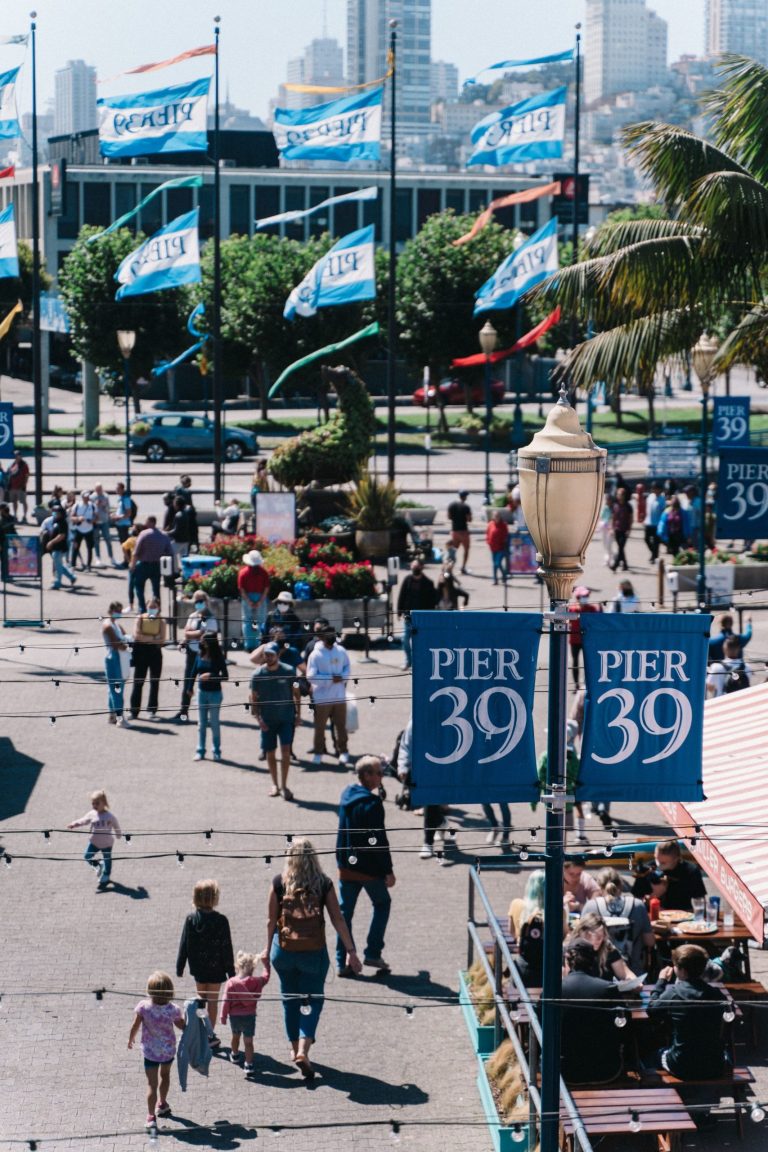 Busy outdoor scene at Pier 39 with people walking and flags waving.